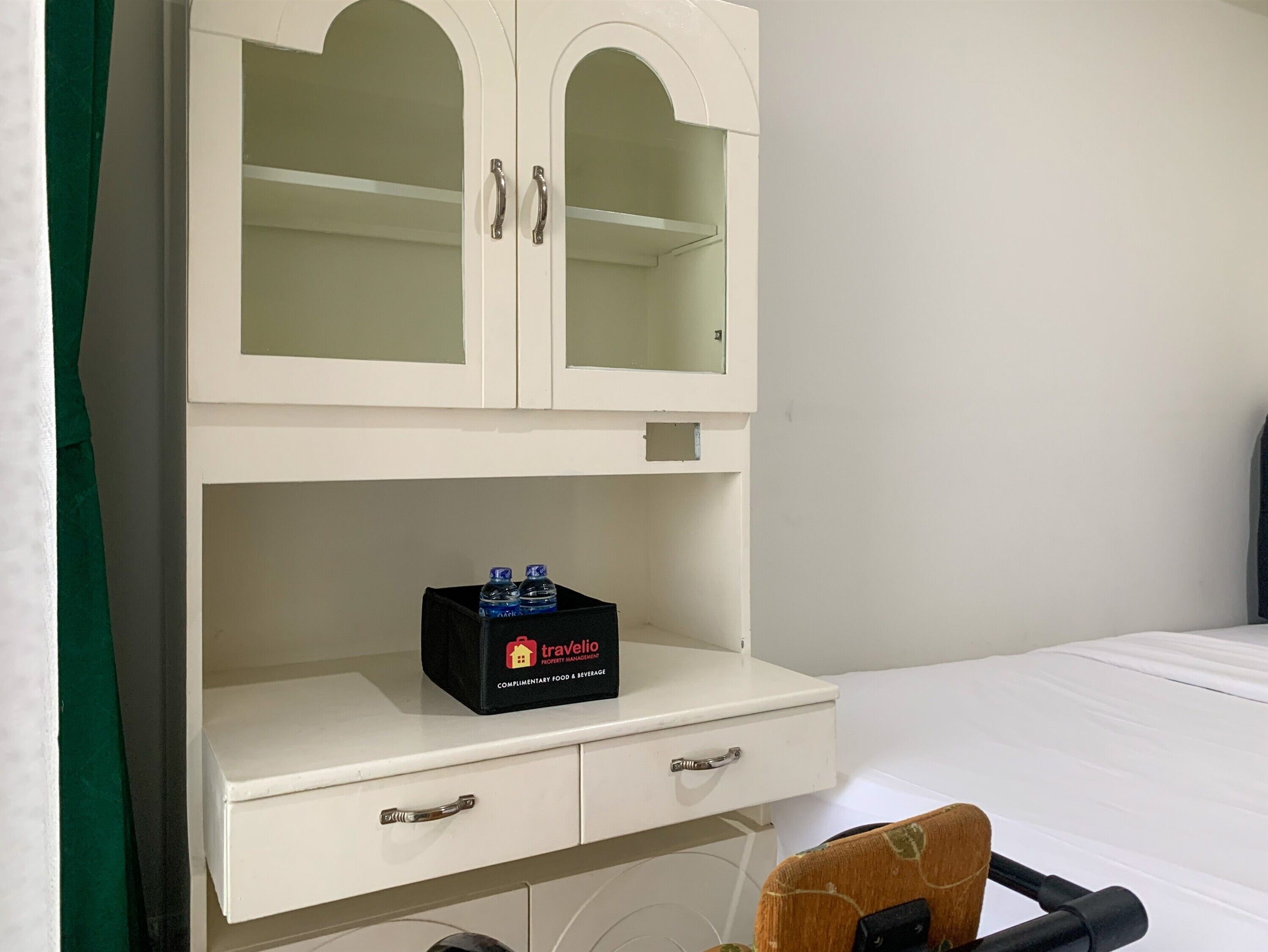 Un meuble blanc à deux portes, une boîte noire portant l’inscription « Travelio » sur l’étagère inférieure, et un lit avec une literie blanche en arrière-plan.