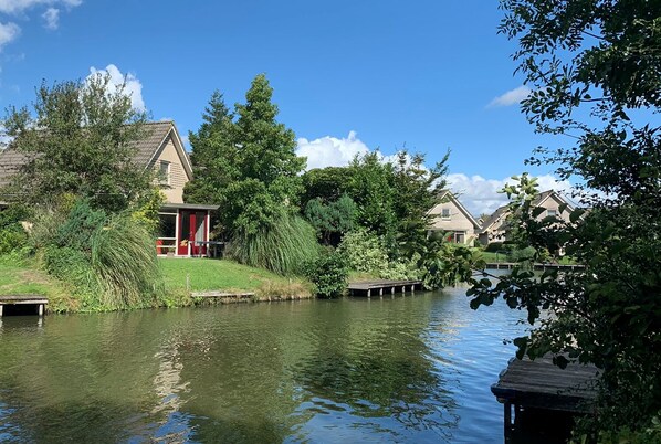 Traditional houses line the serene riverside, surrounded by lush greenery and a wooden dock extending into the calm river.