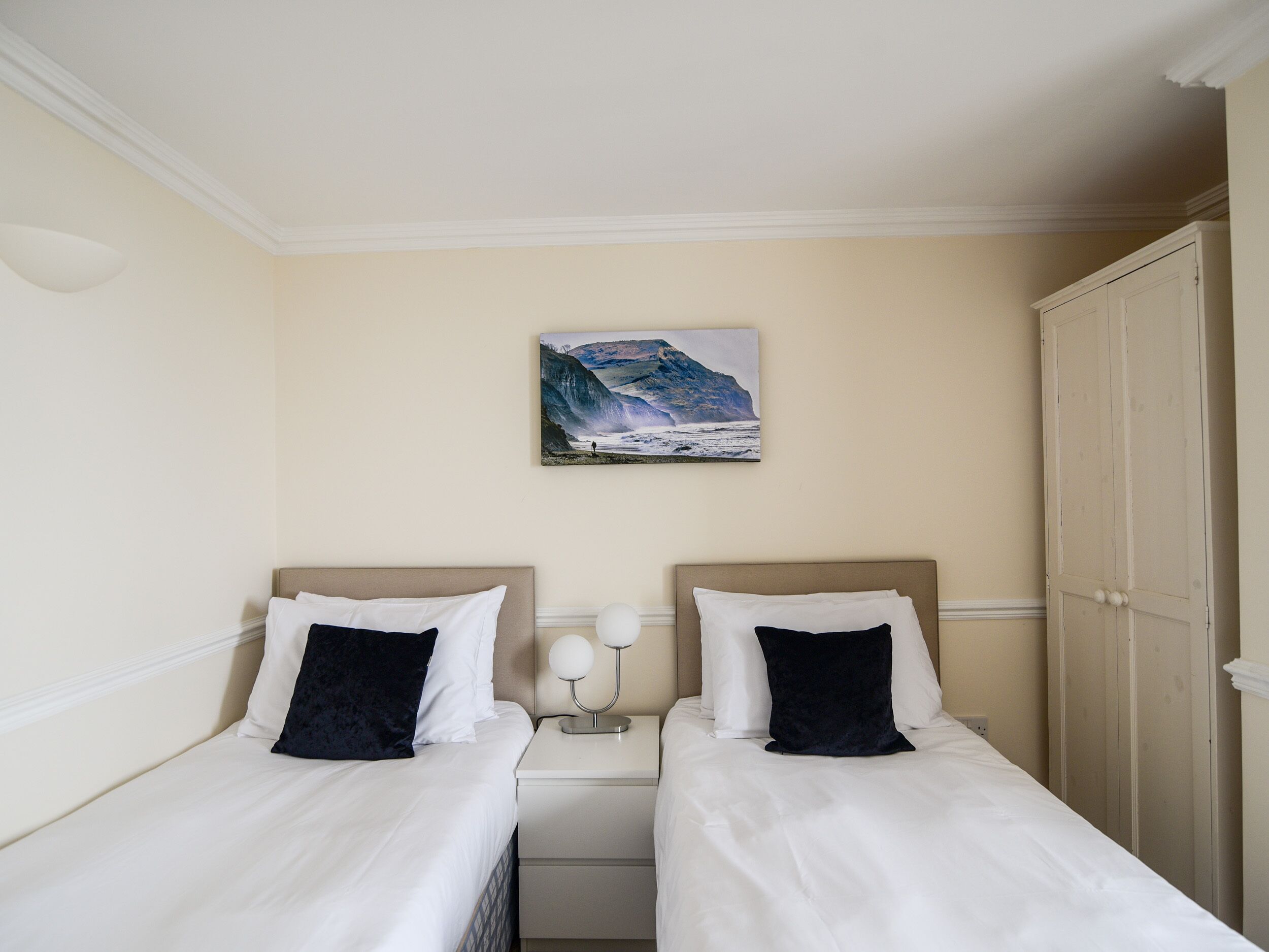 A hotel room with two beds, a nightstand with a lamp, a painting of a mountain landscape, and a wardrobe.