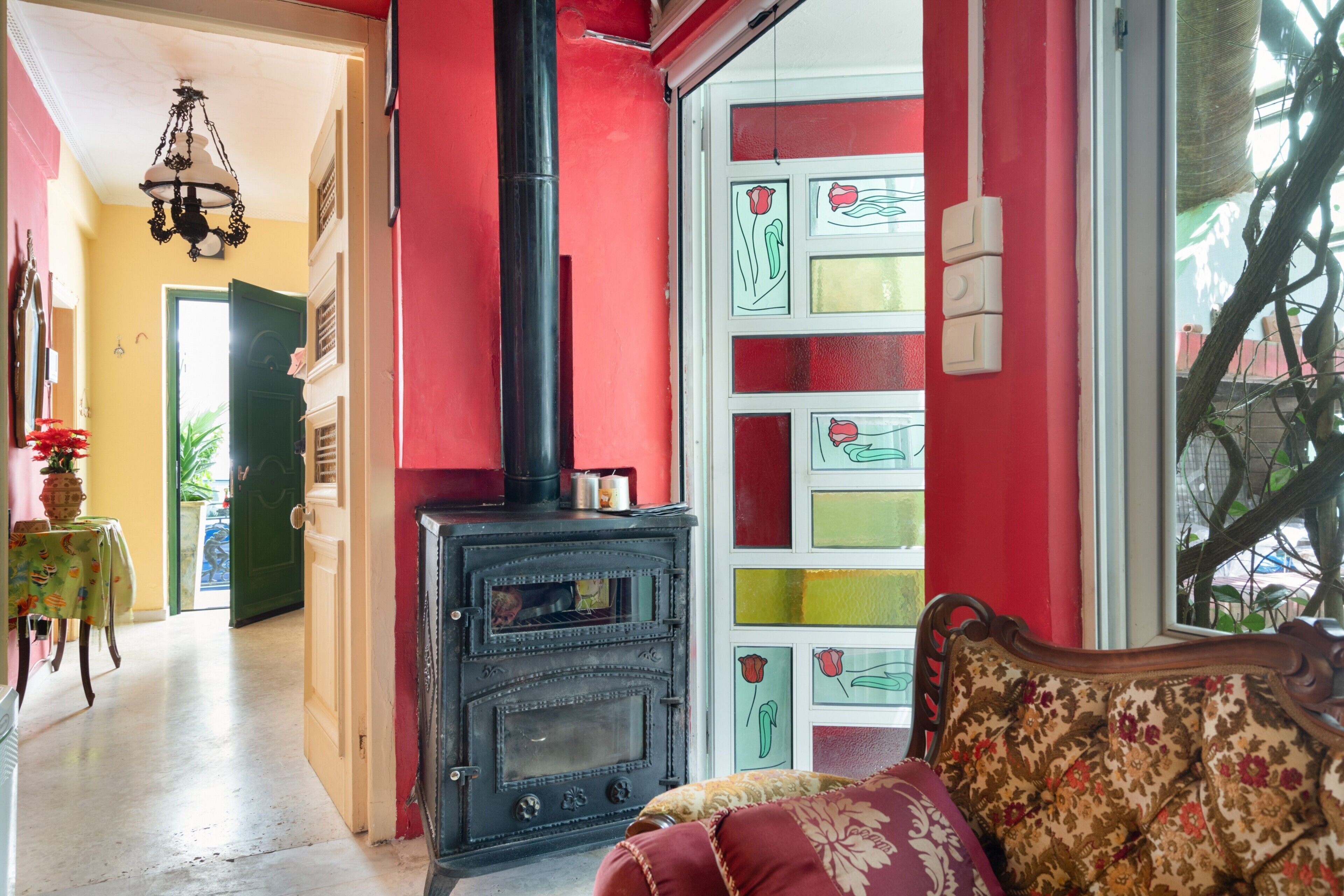 Une pièce avec un mur rouge, un poêle noir, un canapé à motifs et une porte ornée de panneaux de verre colorés.