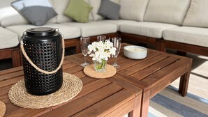 Una mesa de centro de madera con un posavasos tejido, un termo negro con asa de cuerda, un florero con flores blancas y dos vasos.
