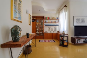 Una sala de estar con una mesa de centro de madera, un jarrón grande, estanterías, un televisor y una alfombra estampada.