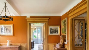 Un couloir avec un banc en bois, une carafe et une statue. Les murs sont ornés de tableaux encadrés et un lustre y est suspendu.
