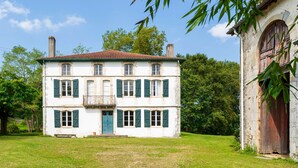 Un bâtiment blanc à deux étages, doté de volets verts, d’un balcon et d’un toit recouvert de tuiles rouges.