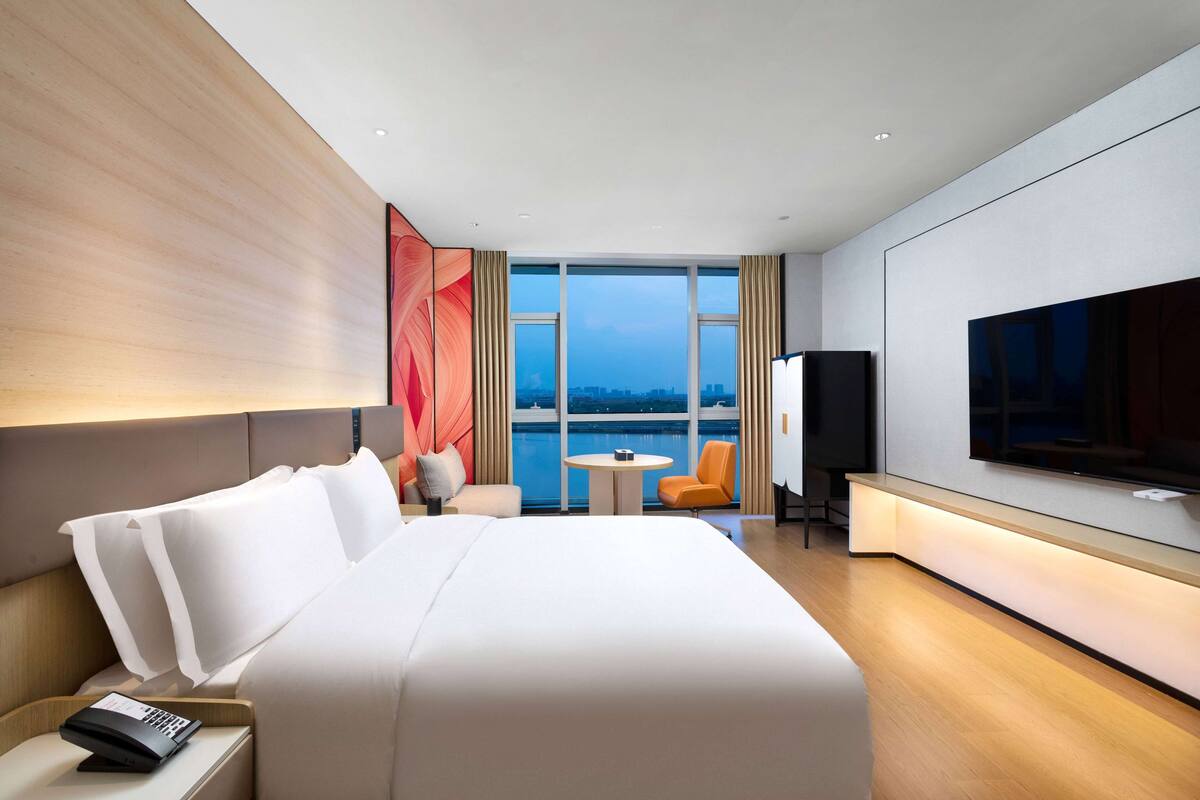 Una habitación de hotel moderna con una cama grande, televisión, una mesita y vistas a la ciudad a través de amplias ventanas.