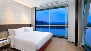 A modern hotel room with a large bed, a nightstand, and a view of a cityscape and water body through floor-to-ceiling windows. - Ramada By Wyndham Huzhou Wuxing (Huzhou)