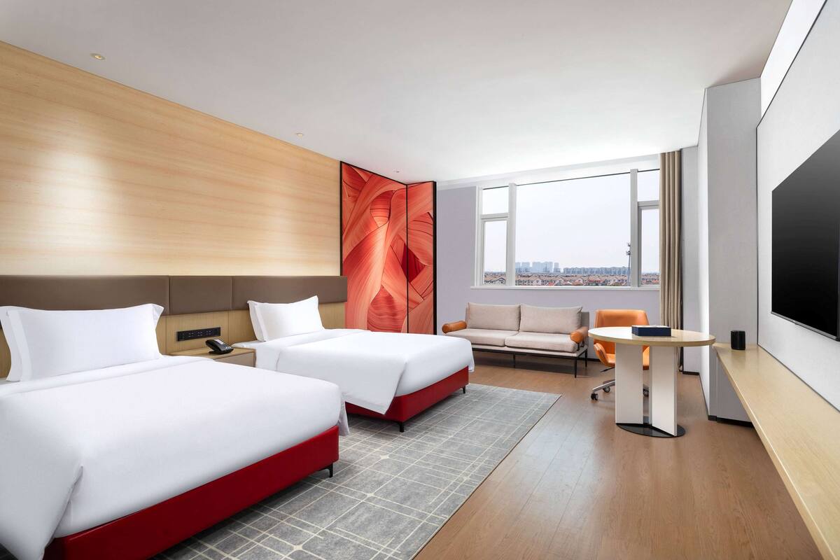 Habitación de hotel con dos camas, un escritorio, una silla, un televisor y un ventanal con vista a la ciudad.