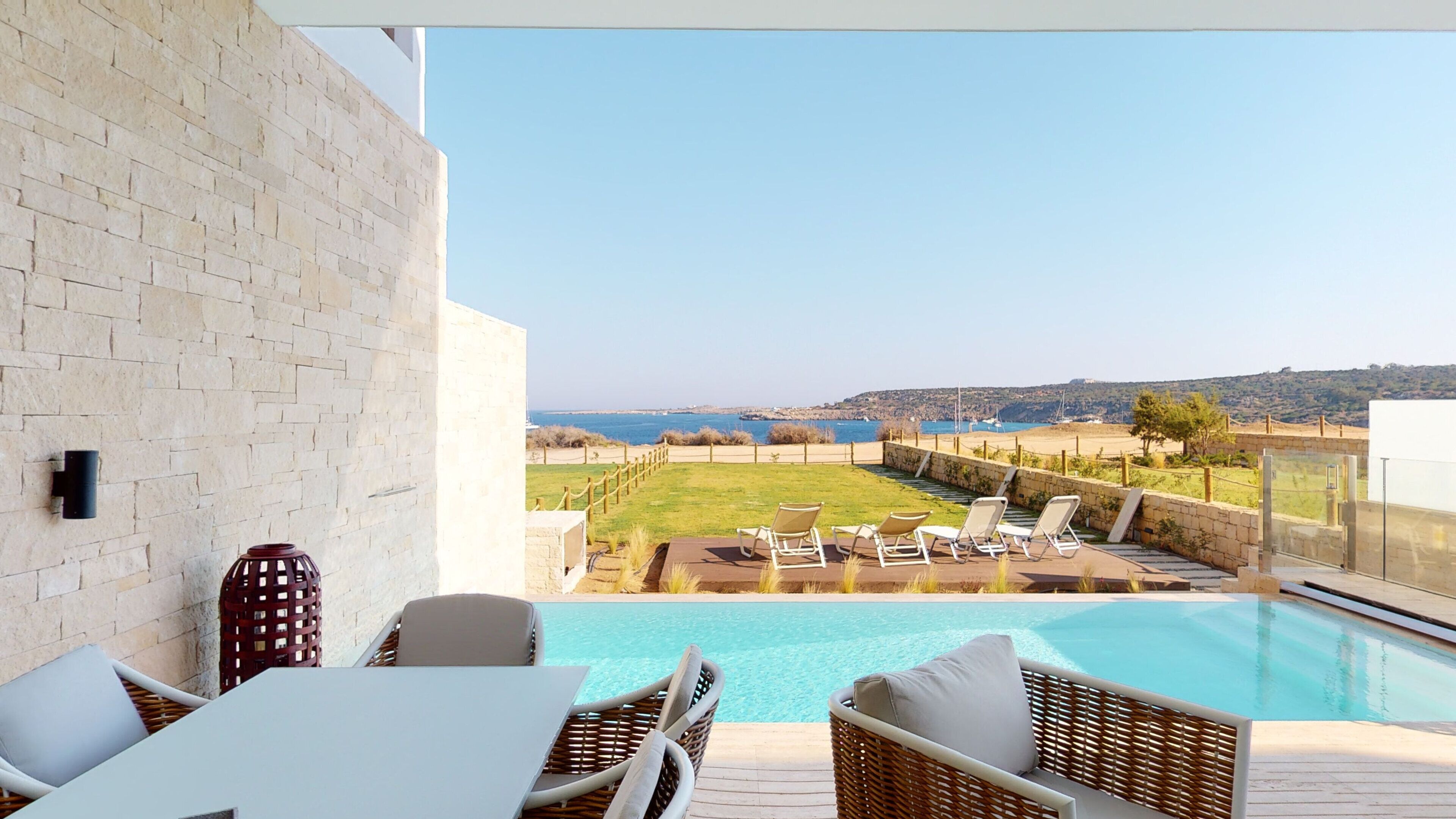 A poolside area with wicker chairs and a table, overlooking a landscaped garden and a view of the sea.