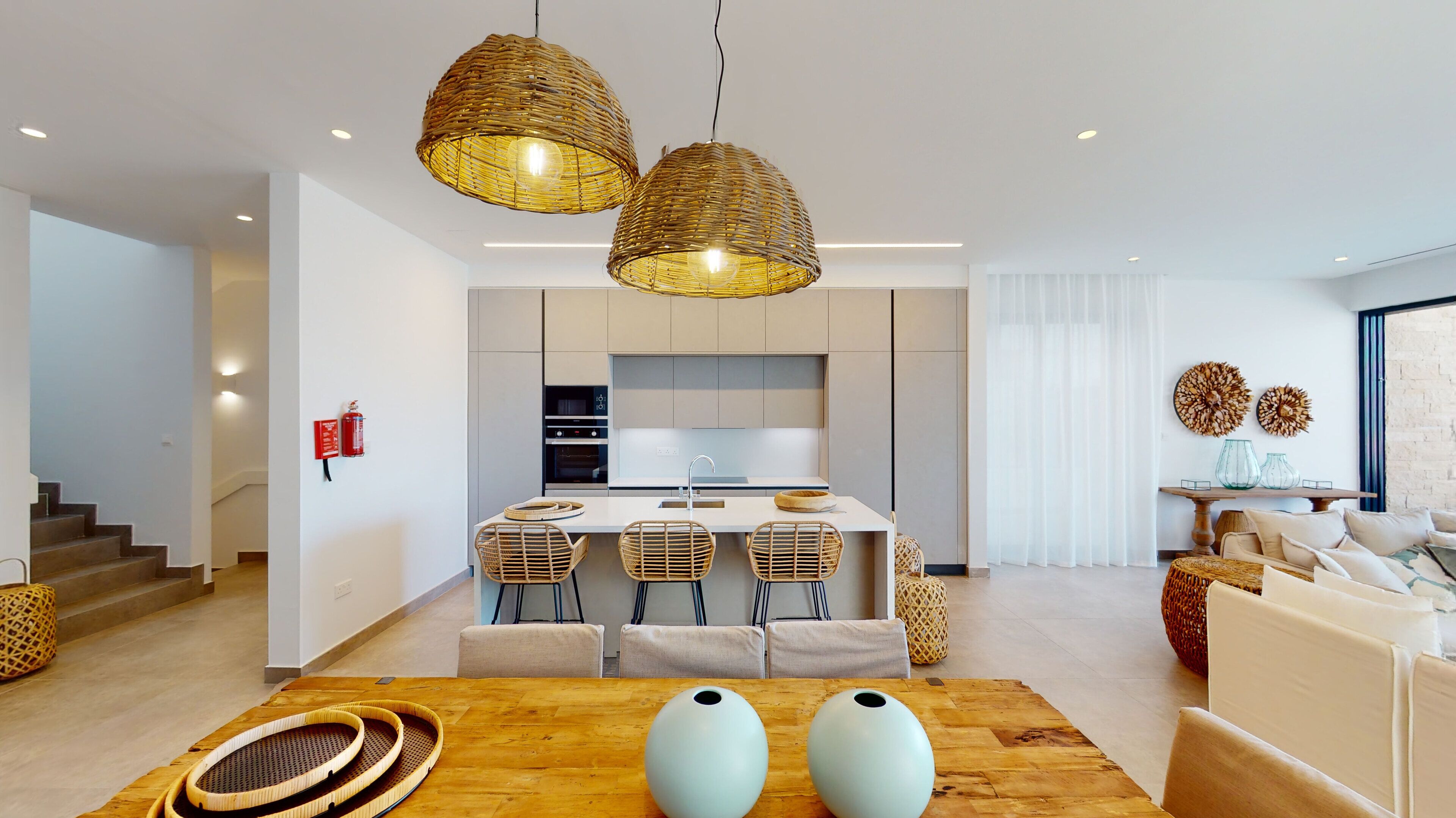 Una cocina moderna con una isla central, dos lámparas colgantes de mimbre y un comedor con una mesa de madera y sillas tejidas.