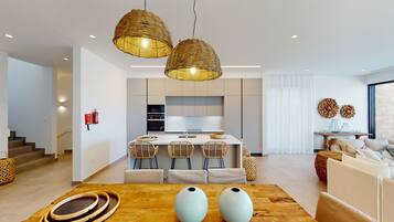 Una cocina moderna con una isla central, dos lámparas colgantes de mimbre y un comedor con una mesa de madera y sillas tejidas.