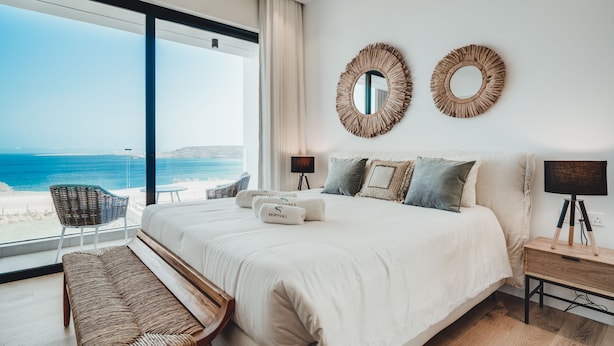 Une chambre avec un grand lit, deux miroirs muraux, une table de chevet avec une lampe, un banc et une vue sur l’océan.