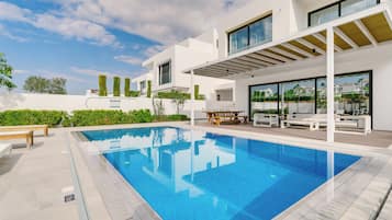 Un área de piscina moderna con una piscina azul cristalina, paredes blancas y una pérgola con asientos.