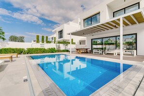 Ett modernt poolområde med en klarblå pool, vita väggar och en pergola med sittplatser.