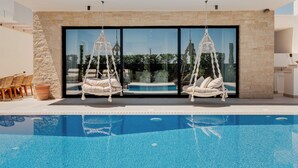 Un espace piscine moderne avec une piscine à l’eau d’un bleu limpide, deux chaises suspendues blanches et un mur en pierre.