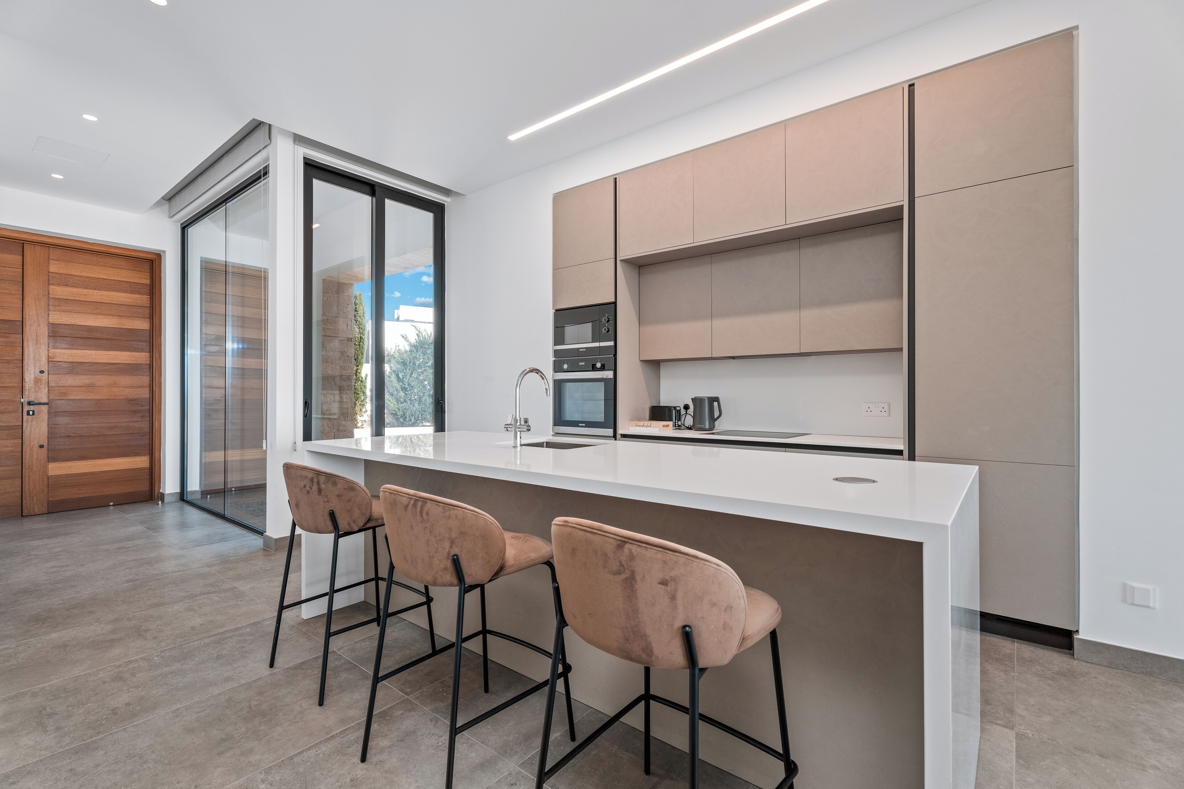 Una cocina moderna con encimera blanca, electrodomésticos de acero inoxidable y un amplio ventanal. Hay tres taburetes de bar y una puerta corrediza de vidrio que da a un patio.