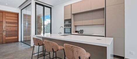 Una cocina moderna con encimera blanca, electrodomésticos de acero inoxidable y un amplio ventanal. Hay tres taburetes de bar y una puerta corrediza de vidrio que da a un patio.