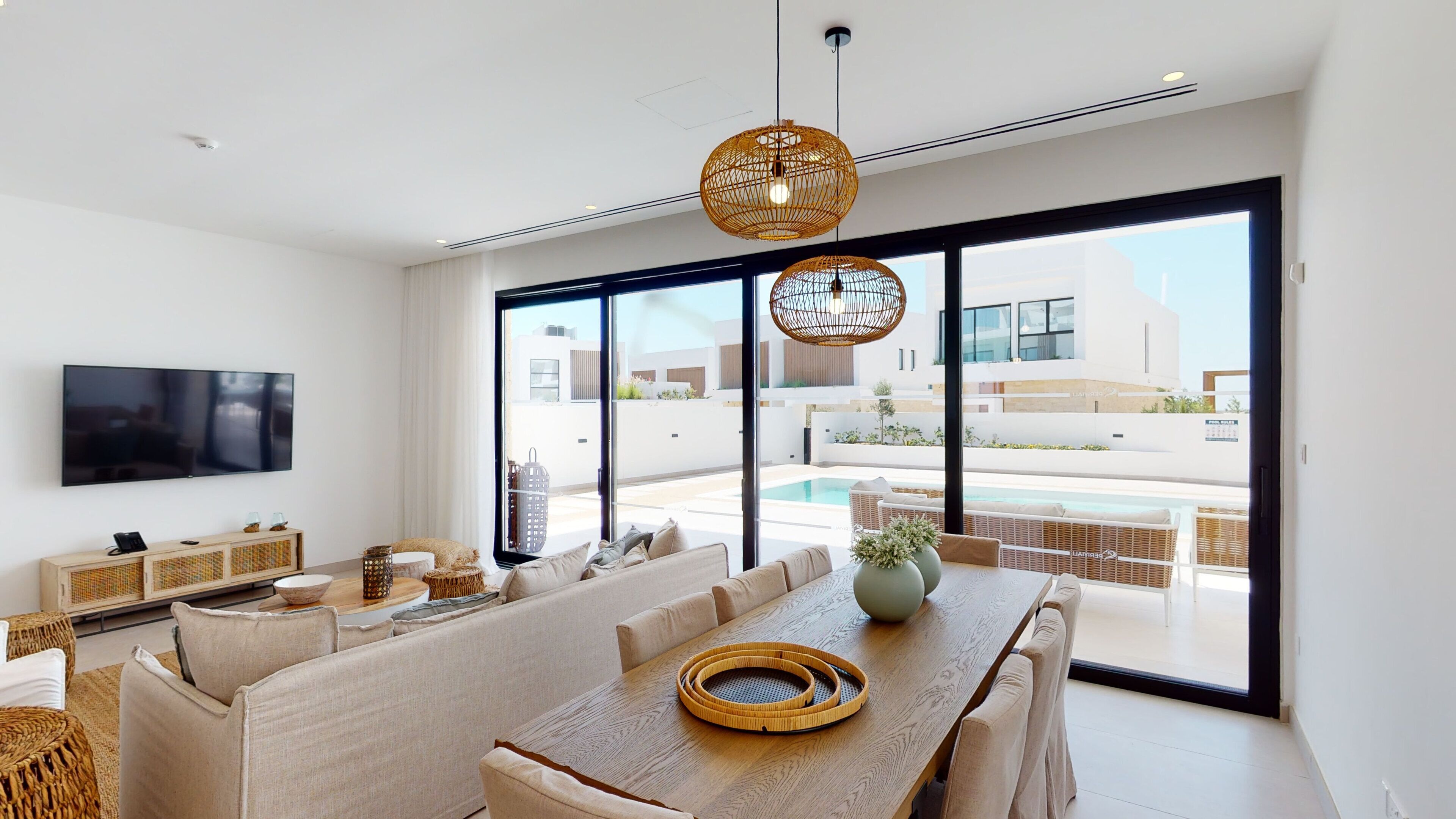 A modern living room with a large TV, comfortable seating, and a view of a pool and buildings.