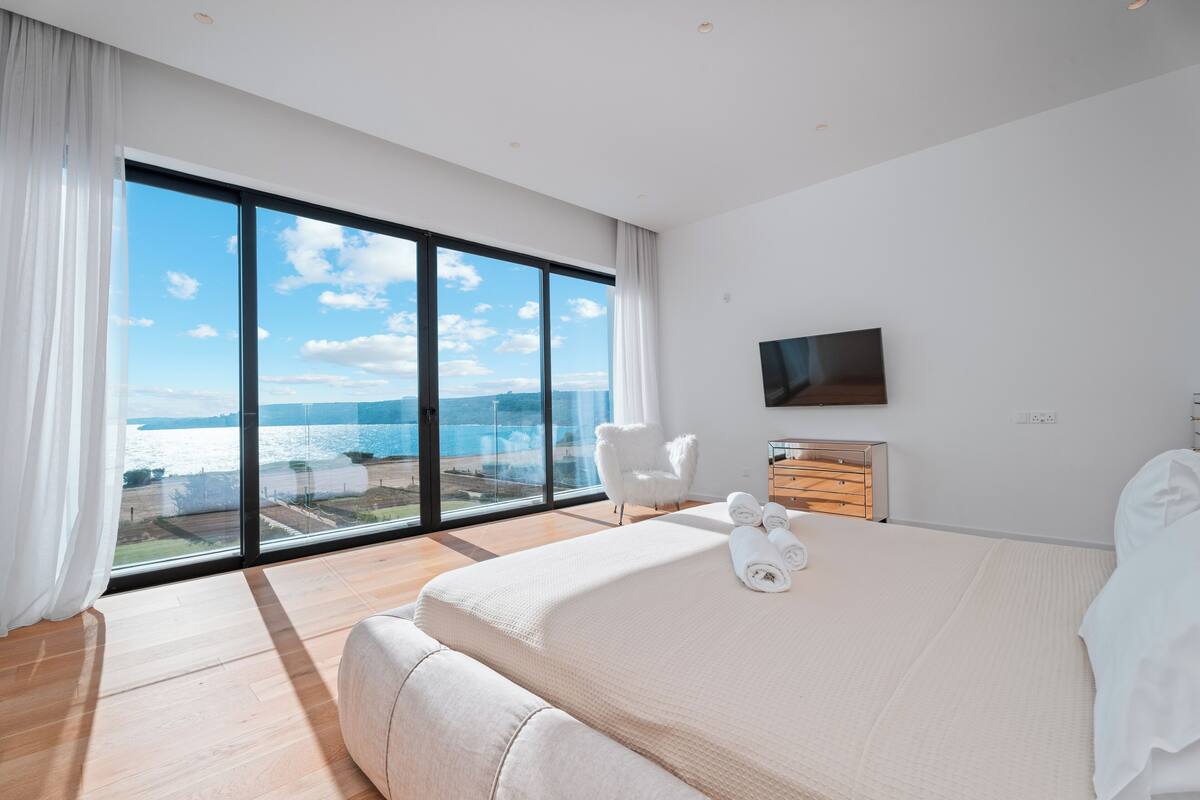 Une chambre d’hôtel moderne dotée d’un grand lit, d’un téléviseur à écran plat, d’une commode en bois et offrant une vue sur la mer grâce à de grandes fenêtres du sol au plafond.