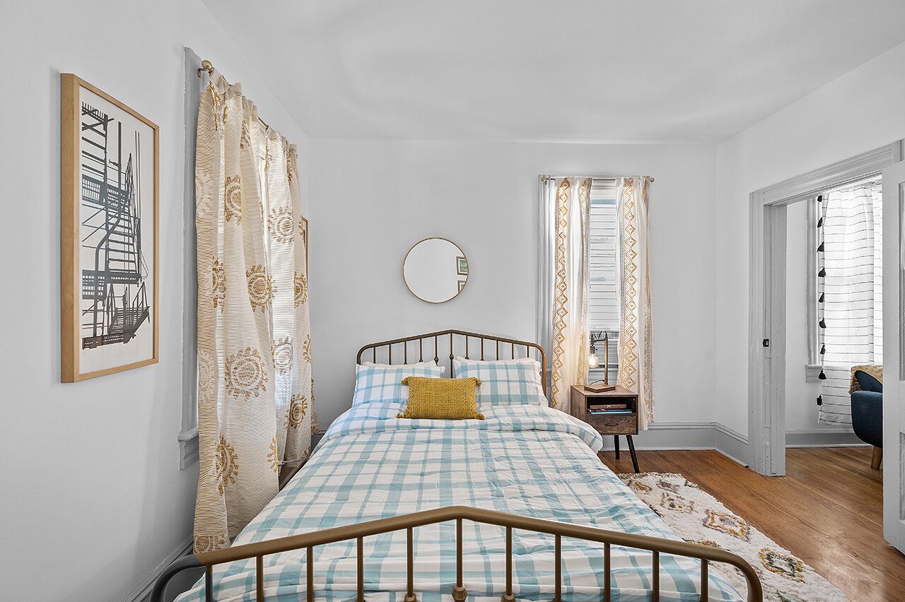 Une chambre à coucher avec un lit, une table de chevet, un miroir et une fenêtre avec des rideaux.
