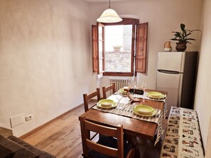 Ein kleiner Essbereich mit einem Holztisch, gedeckt für zwei Personen, einem Fenster mit geöffneten Lamellen und einem Kühlschrank.