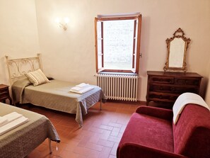 Ein Schlafzimmer mit einem Bett, einem roten Sessel, einem Schminktisch und einem Spiegel, sowie einem Fenster mit Blick auf eine Ziegelwand.