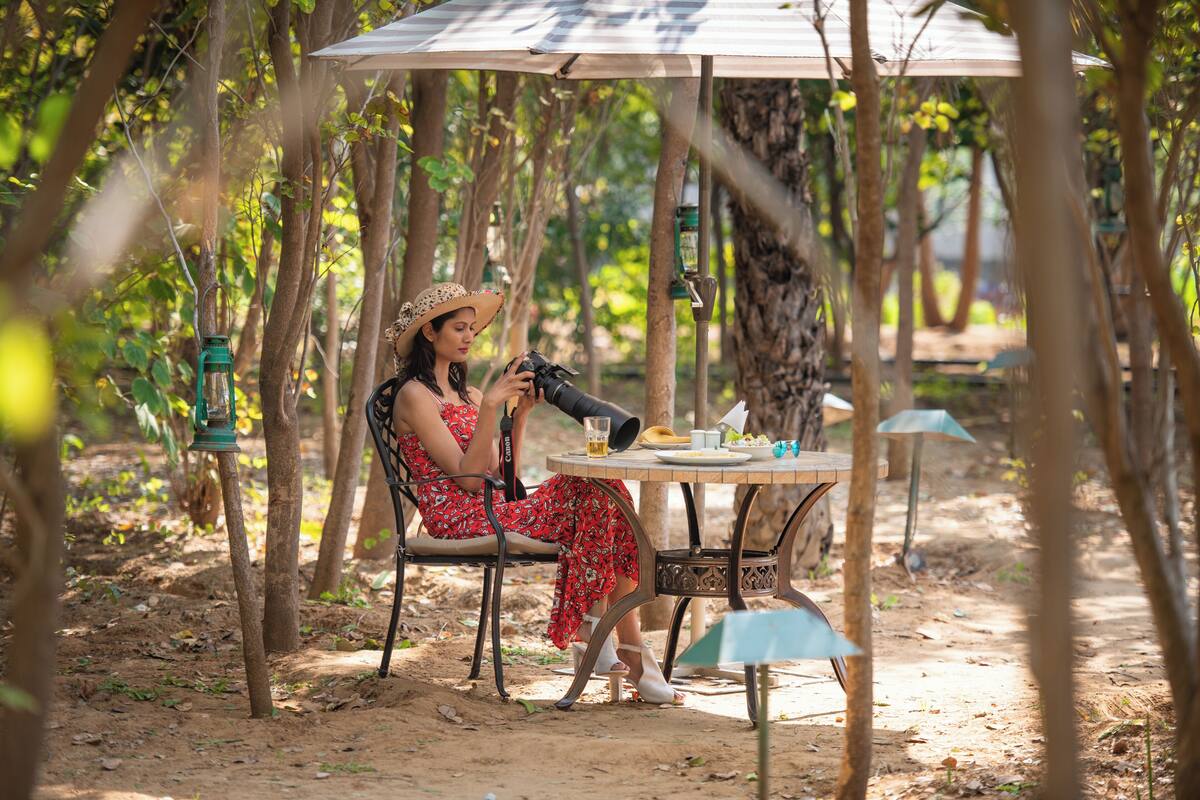 Une personne assise à une table, un appareil photo à portée de main, entourée d’arbres et de mobilier d’extérieur.