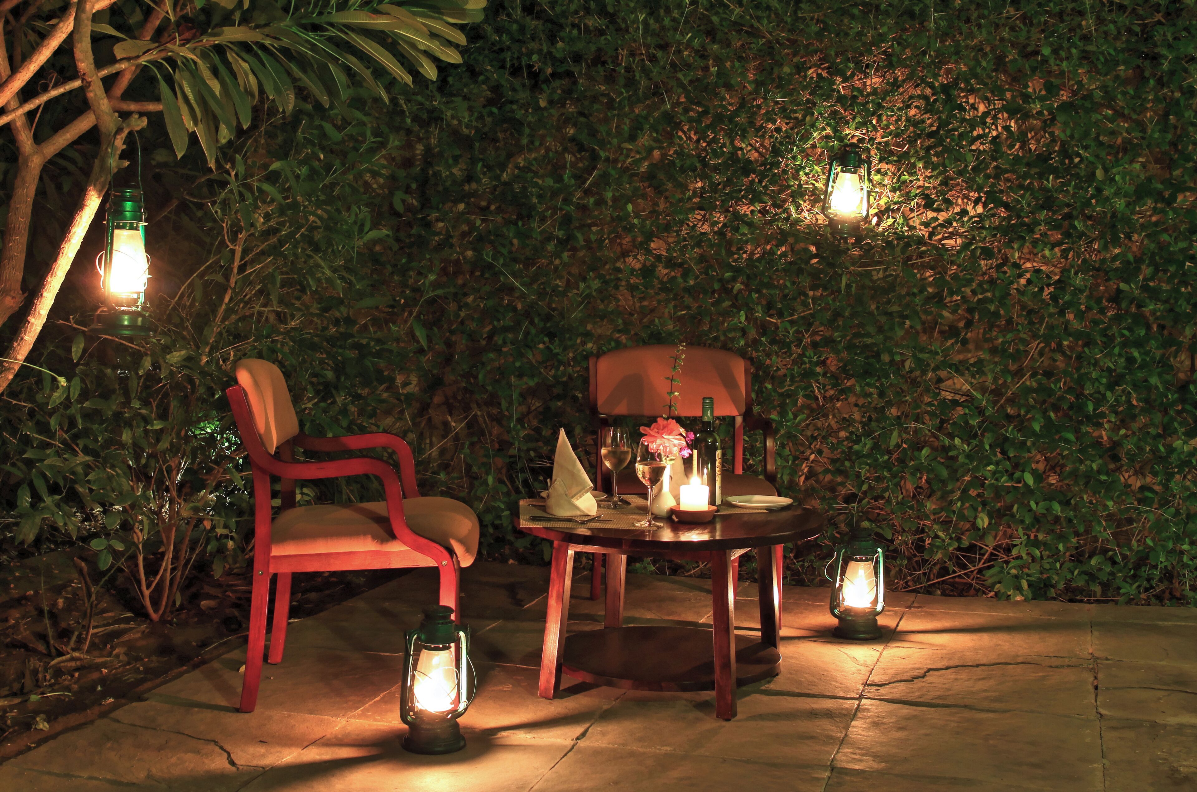 Un espace de restauration en plein air avec une table dressée pour deux personnes, entouré de verdure et illuminé par des lanternes.