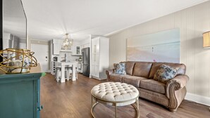 A spacious living area with a brown leather sofa, a round ottoman, and a dining table with chairs. A kitchen with white cabinets and a refrigerator is visible in the background.