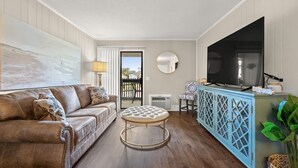 A living room with a brown leather sofa, a round ottoman, a blue cabinet with a TV, and a view through a glass door.