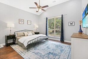 A bedroom with a bed, two bedside tables, a ceiling fan, a window with curtains, and a TV on the wall.