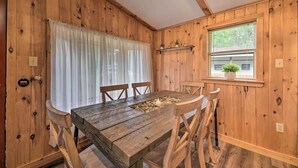 Een houten eettafel met stoelen, een raam met zonwering, en een plank met planten.