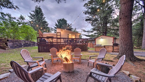 Een vuurkorf omringd door Adirondack stoelen in een bebost achtererf met een houten terras en een klein huisje.