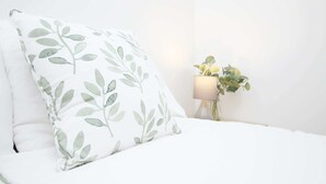 Un lit avec un oreiller blanc orné d’un motif de feuilles vertes, une lampe de chevet et une petite plante en pot, le tout placé devant un mur blanc.