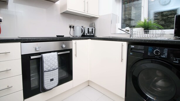 Une cuisine compacte équipée d’un four encastré, d’un lave-linge et d’une fenêtre donnant sur un espace verdoyant.