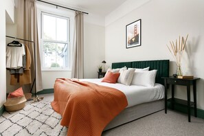 Une chambre à coucher avec un lit, une fenêtre, une table de chevet, une lampe et un tableau encadré au mur.