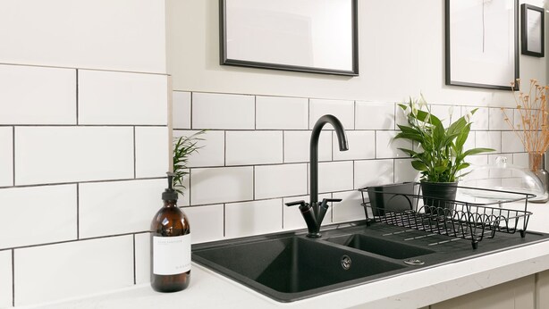 Een keuken met een zwarte kraan, een witte betegelde spatwand en een plant op het aanrecht.