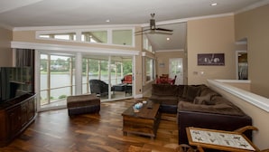A spacious living room with a large sectional sofa, a coffee table, and a television. There is a sliding glass door leading to a balcony with a view of a lake. - Pinehurst Lake House Retreat - Pinehurst Lake Front Property With a Pool (Pinehurst)