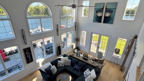A spacious living room with a large sectional sofa, multiple windows, and artwork on the walls. - Huge Cape Cod Style Home on all Sandy beach / Cedar Lake MI (Oscoda)