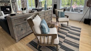 A spacious living room with a fireplace, large windows, and a view of the outdoors. - Huge Cape Cod Style Home on all Sandy beach / Cedar Lake MI (Oscoda)