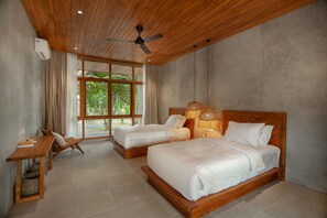Two beds with white bedding, a desk, and a ceiling fan create a serene space with a view of the surrounding greenery.