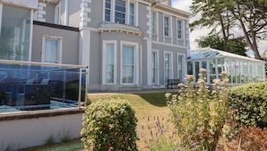Un grand bâtiment à deux étages, doté d’une véranda, entouré de jardins bien entretenus et d’un grand arbre.