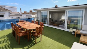 Une terrasse sur le toit avec une table et des chaises en bois, un petit coin salon avec des coussins et une vue sur la mer.