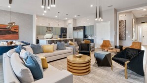 A modern living room with a sofa, armchairs, and a coffee table. There is a kitchen in the background with a refrigerator and cabinets. - Homes by Villatel | Camping Theme BR, Sports Simulator, Theater, Gym, Near Parks (Davenport)
