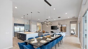 A modern kitchen with a central island, a dining area with blue chairs, and a large TV mounted on the wall. - Homes by Villatel | Camping Theme BR, Sports Simulator, Theater, Gym, Near Parks (Davenport)