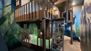 A multi-level play structure with a ladder, a slide, and a climbing wall, set against a mural with forest and mountain imagery. - Homes by Villatel | Camping Theme BR, Sports Simulator, Theater, Gym, Near Parks (Davenport)