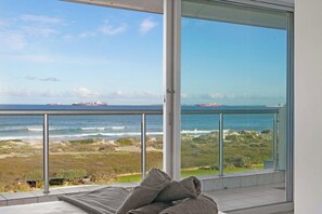 A balcony with a view of the ocean and ships, a bed with pillows, and a glass door. - Sea + Table Mountain Views at dolphin beach (Cape Town)