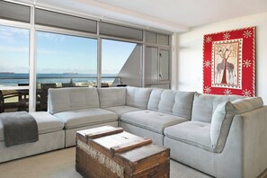 A modern living room with a large sectional sofa, a wooden chest, and a view of the sea through floor-to-ceiling windows.