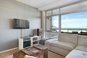 A modern living room with a large window offering a view of the sea and a mountain, a flat-screen TV mounted on the wall, and a cozy sofa.