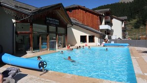 A swimming pool area with people swimming and lounging, a wooden building with a sign, and a mountain in the background. - Resort center, 1st floor, swimming pool, sauna, balcony, tv, ski locker, 50m², Les Orres (Les Orres)