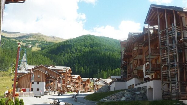 A mountain lodge with wooden architecture, a paved road, and a view of green hills in the background. - Resort center, 1st floor, swimming pool, sauna, balcony, tv, ski locker, 50m², Les Orres (Les Orres)